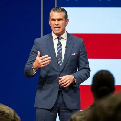 Defense Secretary Pete Hegseth in a navy suit speaks on stage in front of an American flag backdrop, with service members in the foreground