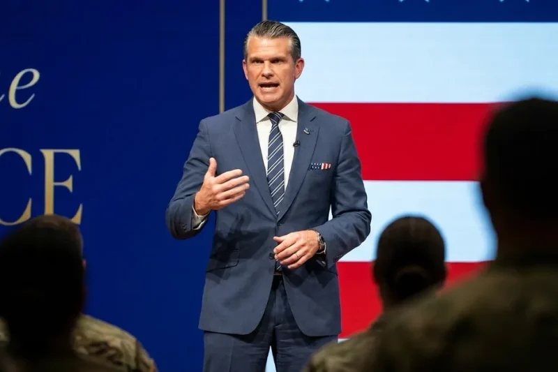 Defense Secretary Pete Hegseth in a navy suit speaks on stage in front of an American flag backdrop, with service members in the foreground