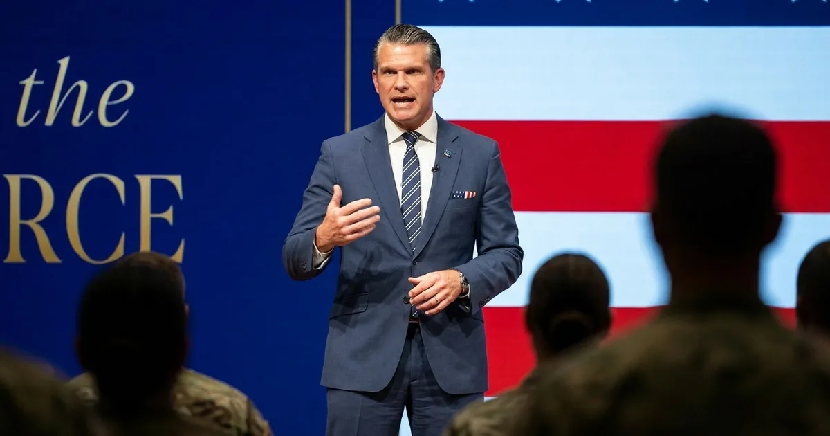 Defense Secretary Pete Hegseth in a navy suit speaks on stage in front of an American flag backdrop, with service members in the foreground
