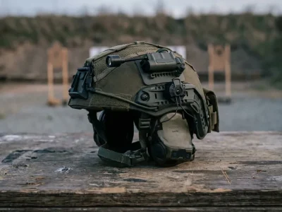 Olive tactical helmet with a rail-mounted electronic headset resting on a wooden table at an outdoor shooting range