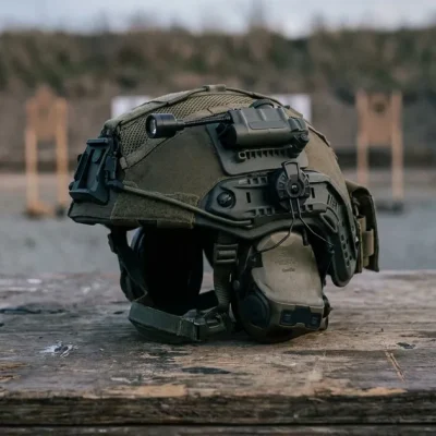 Olive tactical helmet with a rail-mounted electronic headset resting on a wooden table at an outdoor shooting range