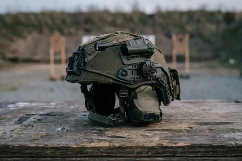 Olive tactical helmet with a rail-mounted electronic headset resting on a wooden table at an outdoor shooting range