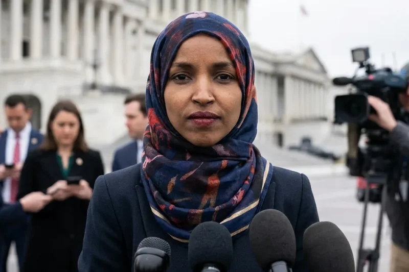 Rep. Ilhan Omar in a navy blazer and patterned hijab speaks into a cluster of microphones outside the US Capitol as reporters and a camera operator look on