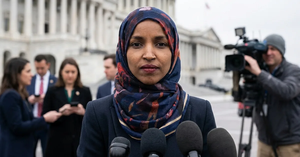 Rep. Ilhan Omar in a navy blazer and patterned hijab speaks into a cluster of microphones outside the US Capitol as reporters and a camera operator look on