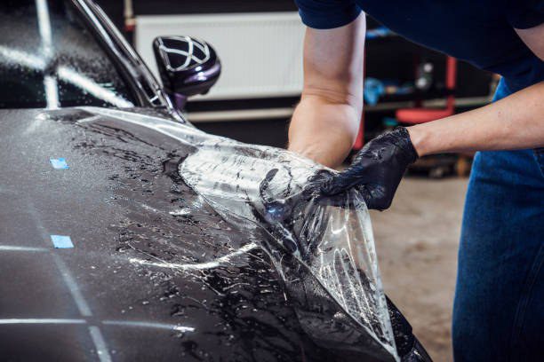 Installer in black gloves smoothing a sheet of clear paint protection film onto a car hood