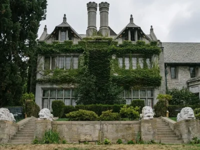 The ivy-covered Gothic-Tudor stone facade of the Playboy Mansion under an overcast sky, with stone lion statues along the terrace wall