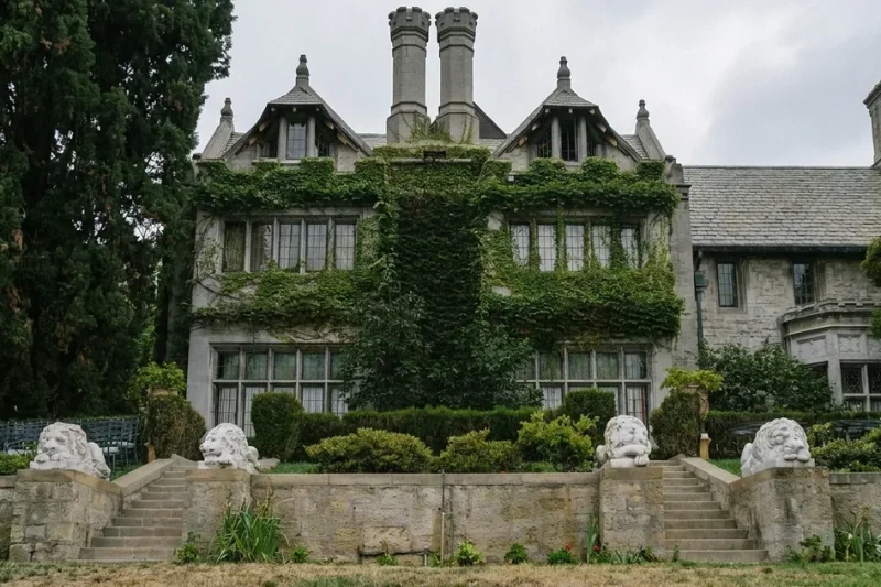 The ivy-covered Gothic-Tudor stone facade of the Playboy Mansion under an overcast sky, with stone lion statues along the terrace wall