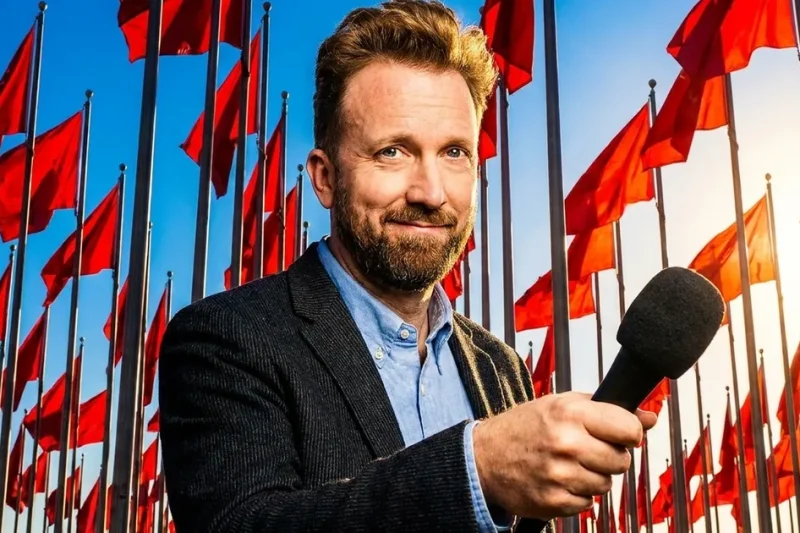 Comedian Jordan Klepper smiling at the camera with a microphone, surrounded by tall red flags against a blue sky