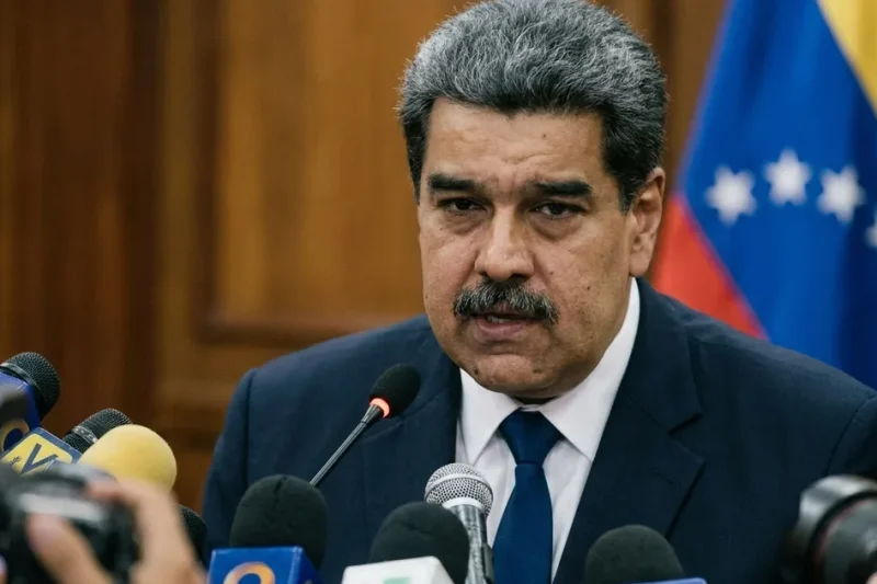 Nicolas Maduro in a dark suit speaking into a cluster of news microphones in front of wood paneling and a Venezuelan flag