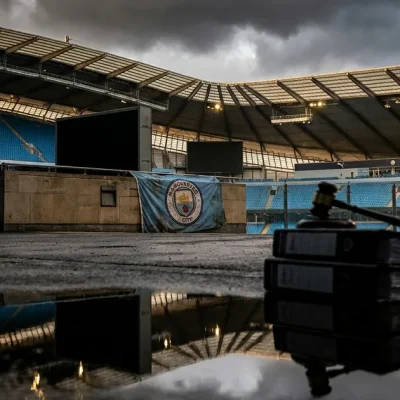 Empty Etihad Stadium stands under storm clouds, a Manchester City banner on the wall, with a stack of legal files and a gavel in the foreground
