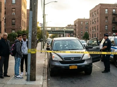 A gray SUV with bullet holes in its windshield behind yellow police tape on a brick-lined Marble Hill street, as neighbors and an NYPD officer look on