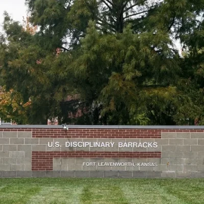 The U.S. Disciplinary Barracks entrance sign on a block-and-brick wall at Fort Leavenworth, Kansas, with trees and a brick building behind it