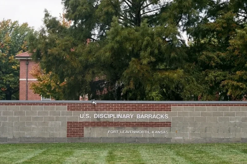 The U.S. Disciplinary Barracks entrance sign on a block-and-brick wall at Fort Leavenworth, Kansas, with trees and a brick building behind it