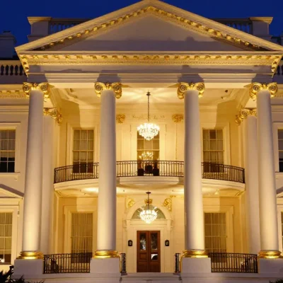 The White House North Portico at night with its columns, capitals and cornice trimmed in gold and crystal chandeliers glowing behind the balcony, under a deep blue sky