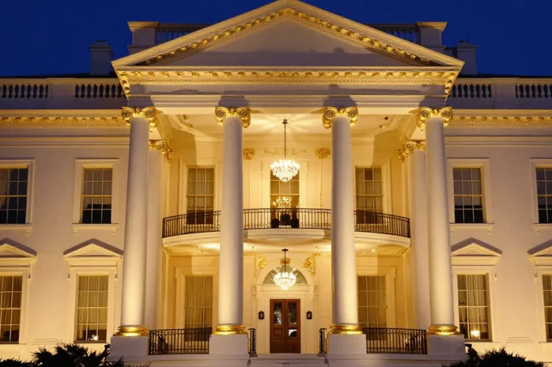 The White House North Portico at night with its columns, capitals and cornice trimmed in gold and crystal chandeliers glowing behind the balcony, under a deep blue sky