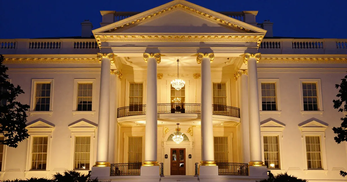 The White House North Portico at night with its columns, capitals and cornice trimmed in gold and crystal chandeliers glowing behind the balcony, under a deep blue sky