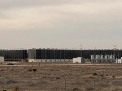 A large dark data center building with cooling units and power lines on flat plains behind a chain-link fence under an overcast sky