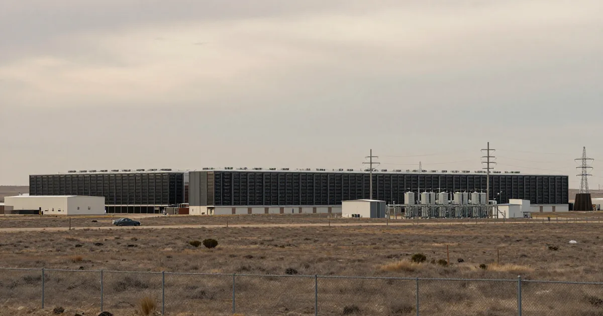 A large dark data center building with cooling units and power lines on flat plains behind a chain-link fence under an overcast sky