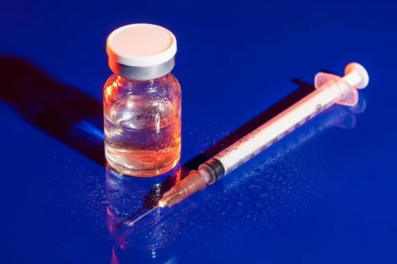 An unlabeled glass vial with a white cap and an insulin syringe lying on a wet cobalt blue surface under hard orange light