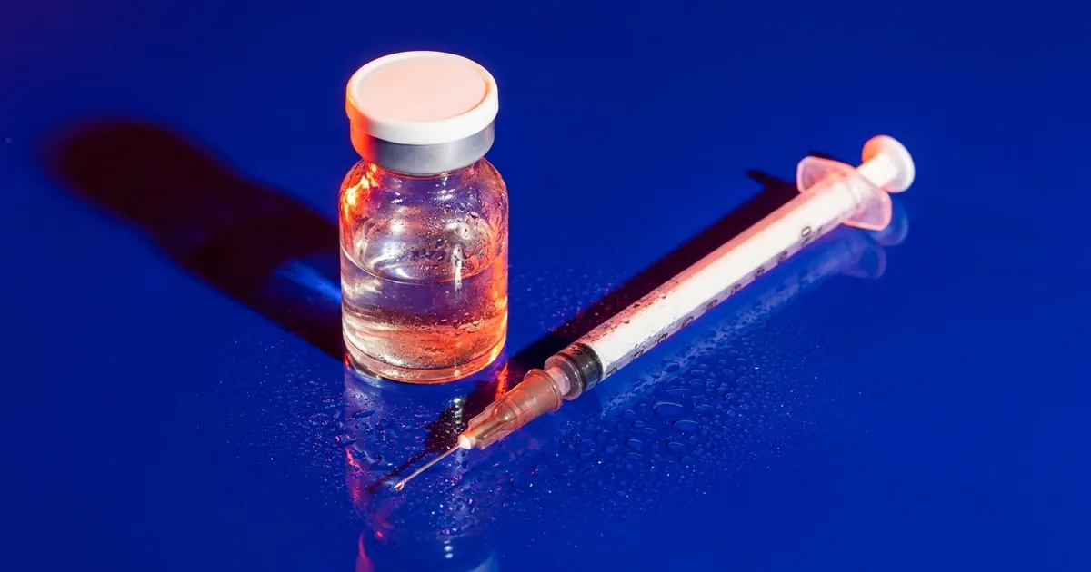An unlabeled glass vial with a white cap and an insulin syringe lying on a wet cobalt blue surface under hard orange light