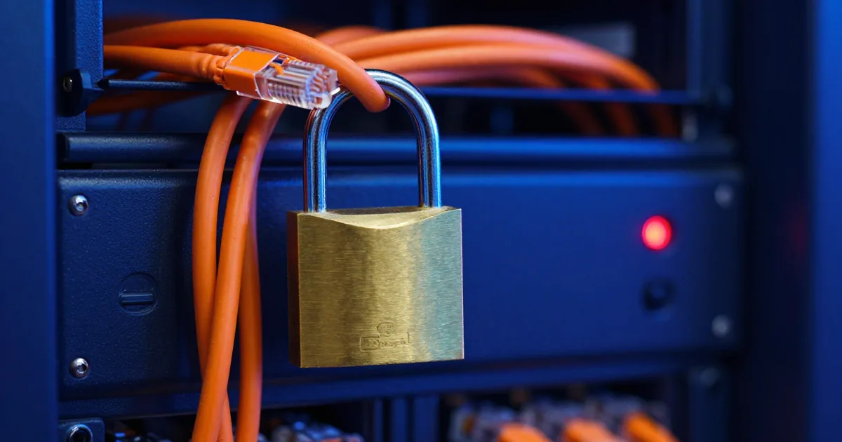 Brass padlock hanging from orange network cables in front of a server rack with a red warning light