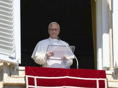 Pope Leo XIV, in white, stands behind a clear lectern at the open window of the Apostolic Palace above a red banner.