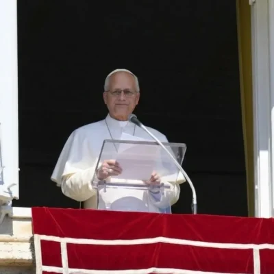 Pope Leo XIV, in white, stands behind a clear lectern at the open window of the Apostolic Palace above a red banner.