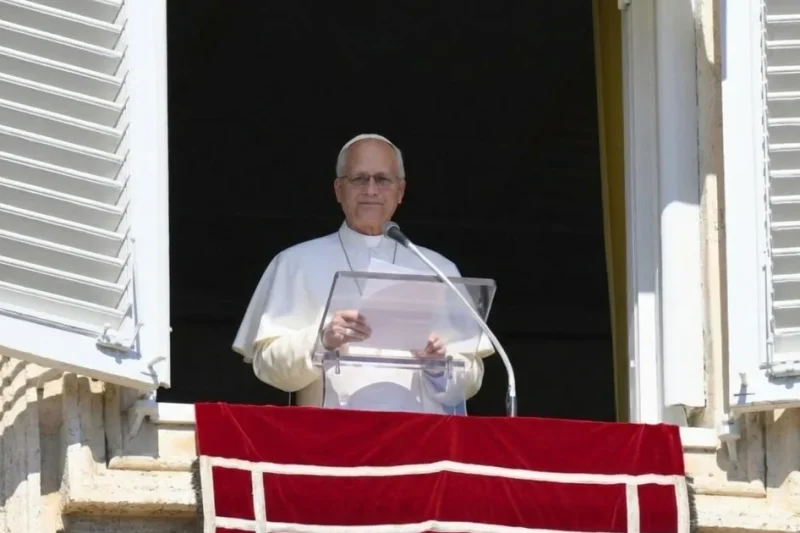 Pope Leo XIV, in white, stands behind a clear lectern at the open window of the Apostolic Palace above a red banner.