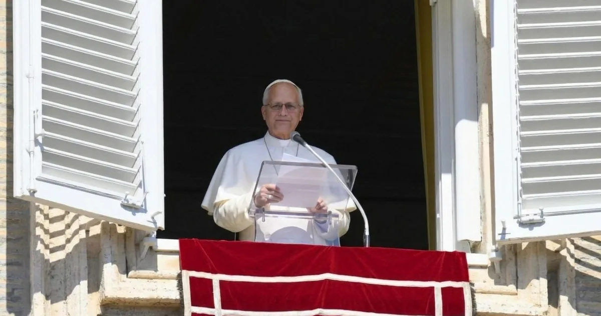 Pope Leo XIV, in white, stands behind a clear lectern at the open window of the Apostolic Palace above a red banner.