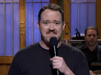 Shane Gillis in a black T-shirt holds a microphone on the Saturday Night Live stage with a sheepish expression.