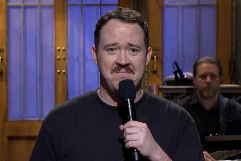 Shane Gillis in a black T-shirt holds a microphone on the Saturday Night Live stage with a sheepish expression.