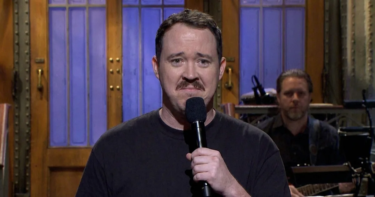 Shane Gillis in a black T-shirt holds a microphone on the Saturday Night Live stage with a sheepish expression.