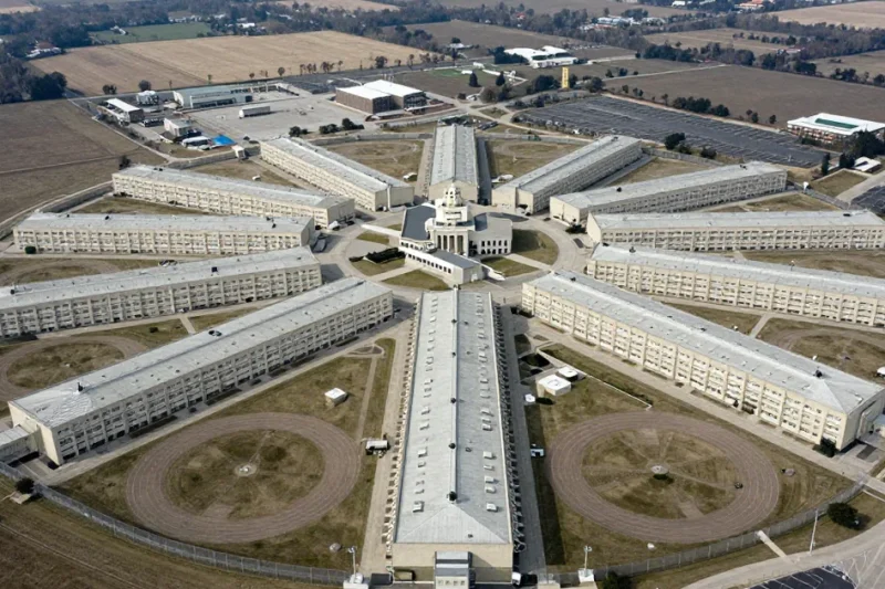 Aerial view of a sprawling federal prison complex with radiating cellblock wings, recreation yards and perimeter fencing surrounded by farmland