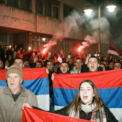 Supporters holding red, blue and white Serbian flags cheer at night outside a building as flares burn behind them