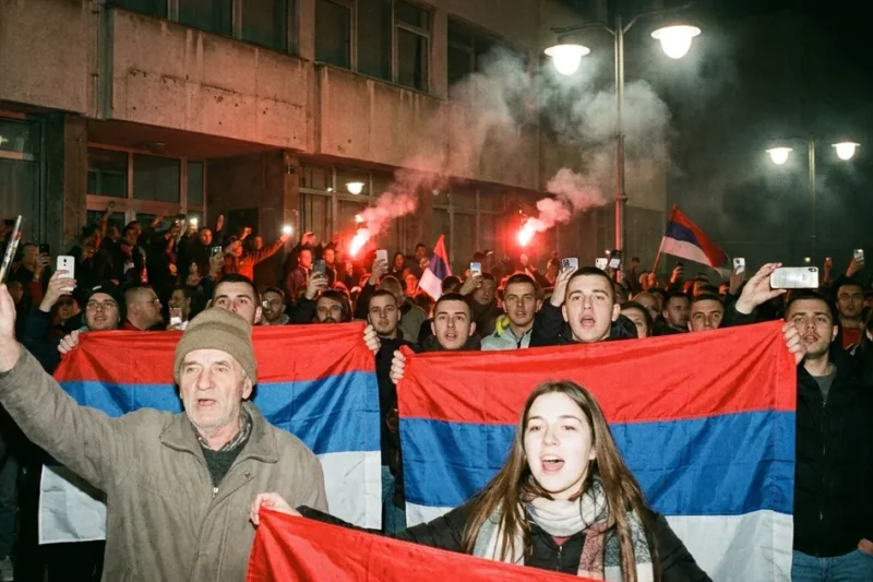 Supporters holding red, blue and white Serbian flags cheer at night outside a building as flares burn behind them