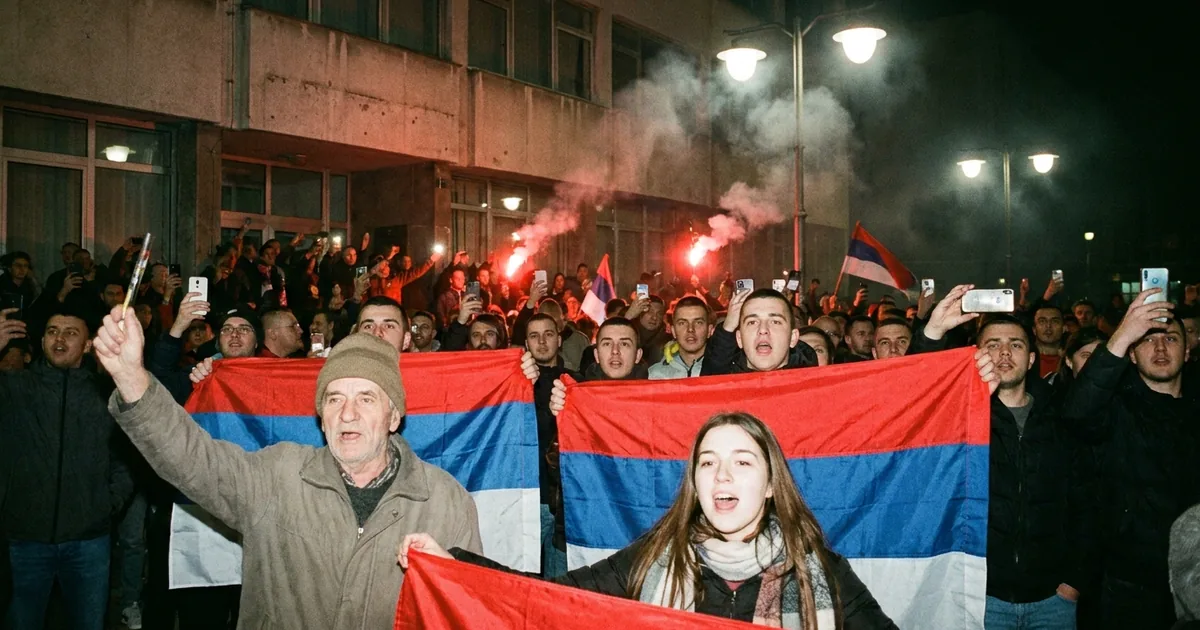 Supporters holding red, blue and white Serbian flags cheer at night outside a building as flares burn behind them
