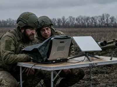 Two Ukrainian soldiers in helmets crouch at a folding table in a muddy field, looking at a laptop beside a flat satellite terminal and a small interceptor drone launcher