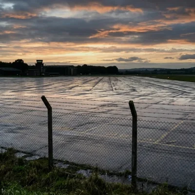 A wet, empty aircraft apron behind a security fence at dusk with a single vehicle in the distance