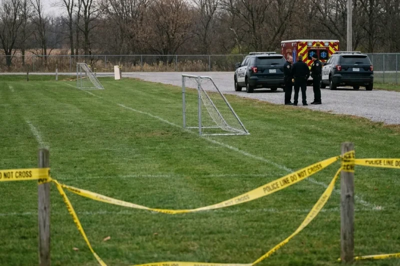 Illustrative scene: police tape across a mown youth soccer field, with patrol SUVs and an ambulance parked on a gravel road beyond the goals